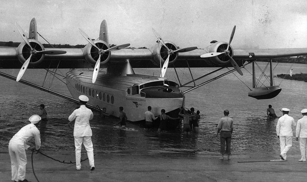 A Sikorsky S-42 landing and pulling into port. The plane has four large motors mounted on the wing that stretched over the body of the craft. The plane is painted in silver and black. The plane is surrounded by workers in naval uniform guiding the plane in to port. There are also many people in swimming suits looking on or pushing the plane.
