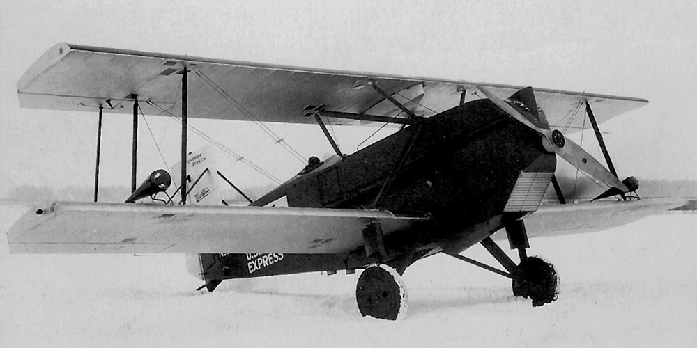 A Curtiss Carrier Pigeon sits in a snow covered field. The plane is marked as owned by the National Air Transport Company. The biplane has a mostly metal body and is equipped with lights on the wings for night flights.