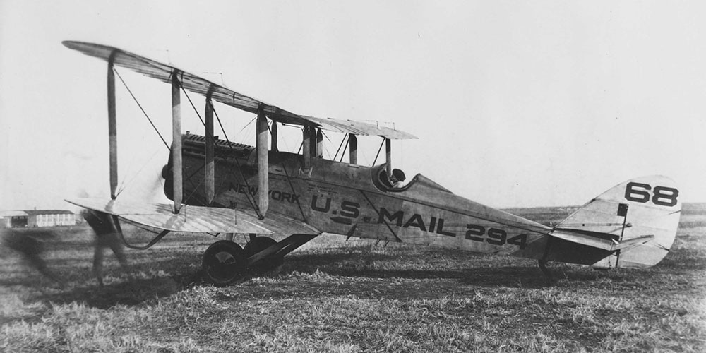 A DeHavilland DH-4B, designated #294 sits in a field getting ready for takeoff. The pilot sits in the cockpit while two men run out of frame as they start the propeller. In the far back is the hanger for the airfield from which the pilot is taking off.