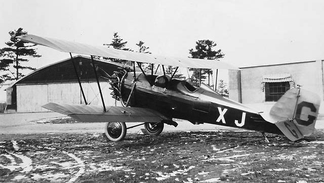 The picture is of a Pitcairn PA-7S. The plane has the logo of the Royal Canadian Air Force with the designation XJ painted on the side. The plane is on a muddy field in front of an airport.