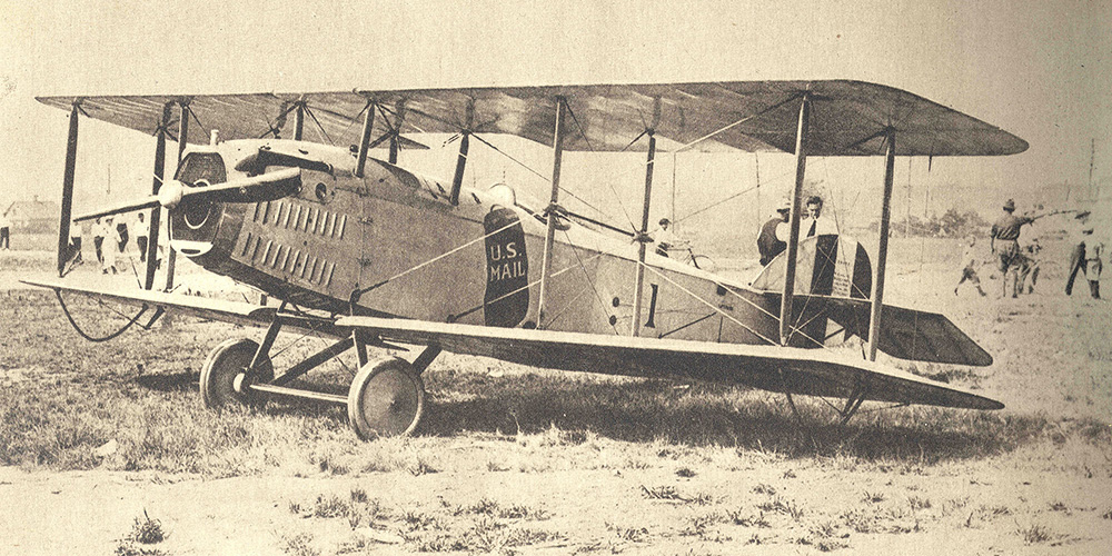 A Standard JR-1B biplane on a field. There are as many as ten behind the plane on the field. The plane has a mail bag painted on its side that reads US Mail along with the number 1.