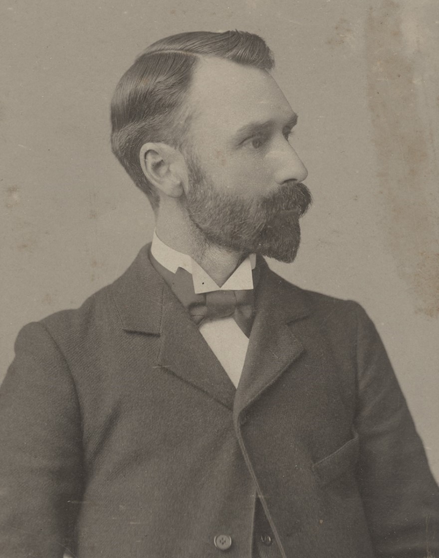 Black and white photographic portrait of a man from the shoulders up, in a suit with bow tie. He has a full, yet manicured beard and mustache and has his head turned to the side.