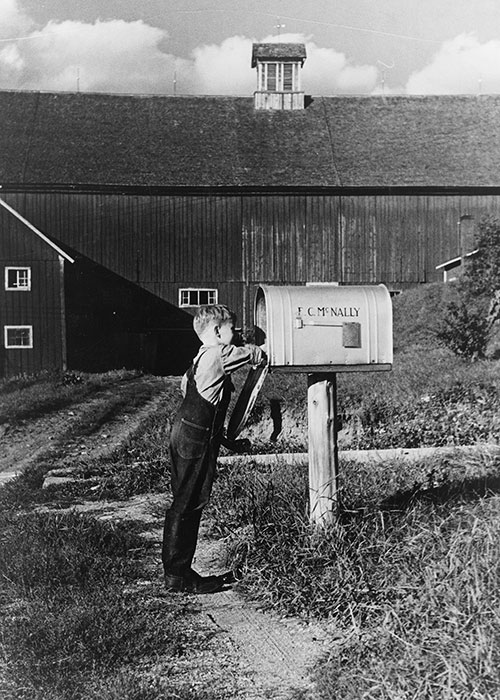 A young boy in overalls reaches into a mailbox bearing the name F.C. McNally.
