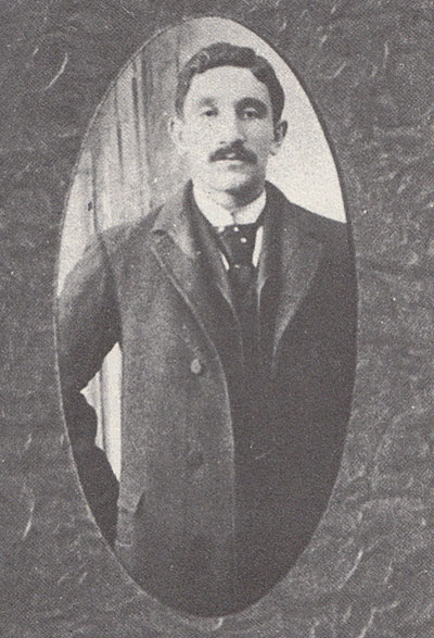 A black and white portrait of a young man. He is standing, and he wears an overcoat. He has a mustache, short hair, and a neutral expression. His gazes faces the camera.