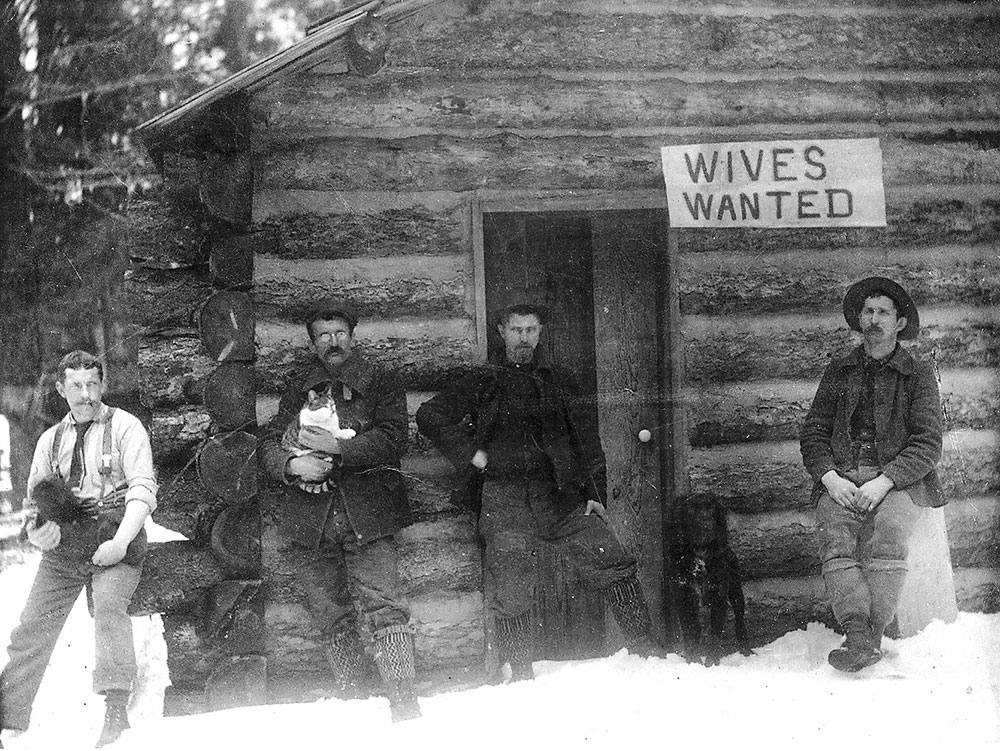 A black-and-white photograph of four men standing on snow-covered ground in front of a log cabin. Affixed to the cabin is a sign that reads, Wives Wanted.