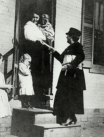 A female mail carrier on steps handing a letter to a woman who is in a doorway holding a child.